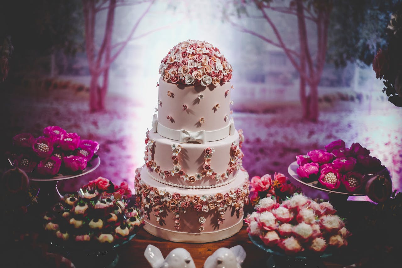 Beautifully decorated wedding cake with floral design surrounded by desserts at an indoor event.